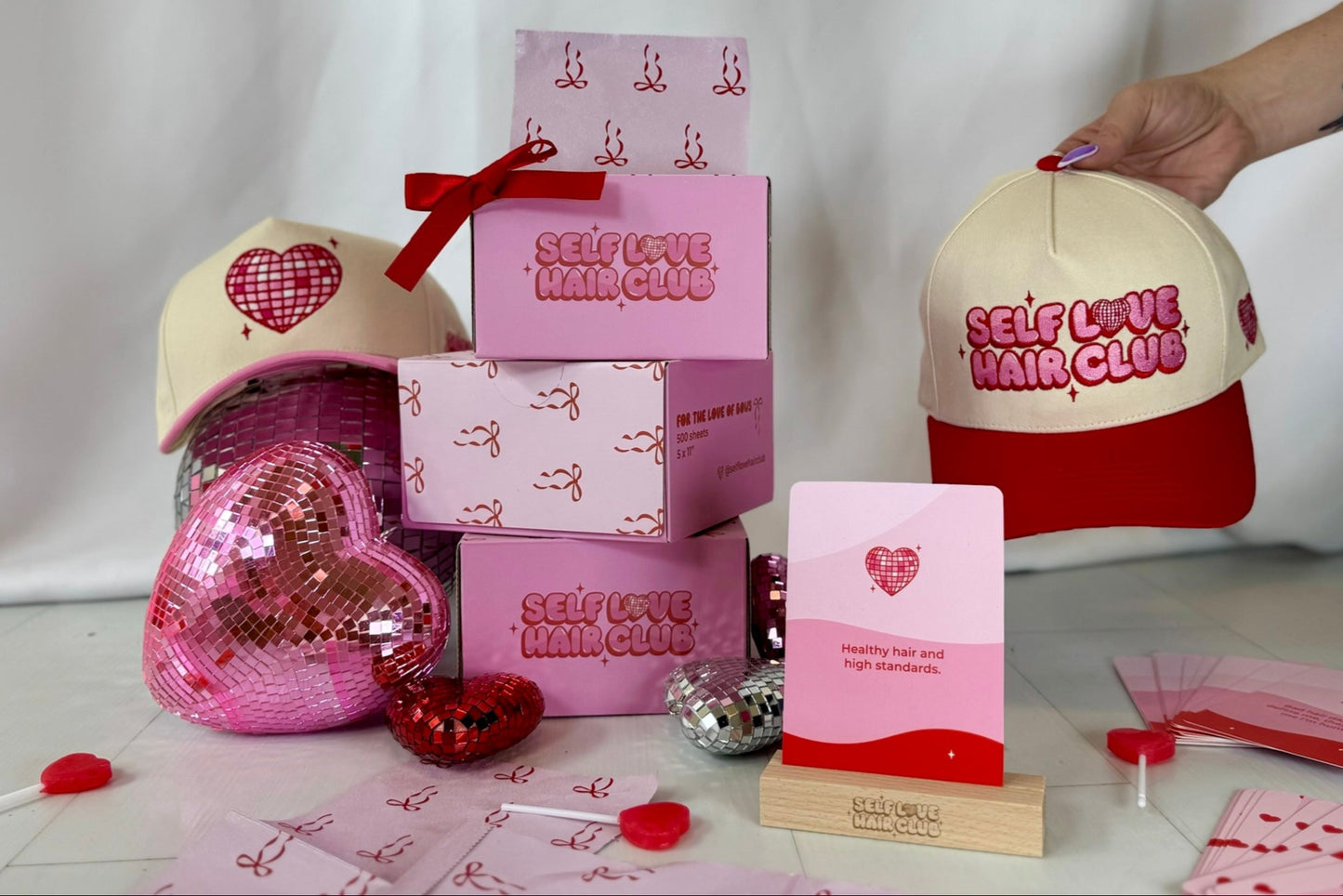 Self Love Hair Club product display featuring foil boxes with bow design, two trucker hats, affirmation card on wooden stand, and heart-shaped disco balls with lollipops.