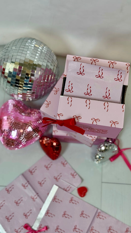 Close up of Self Love Hair Club foil boxes with red bow design, surrounded by silver and heart shaped disco balls, and red lollipops on a white surface.