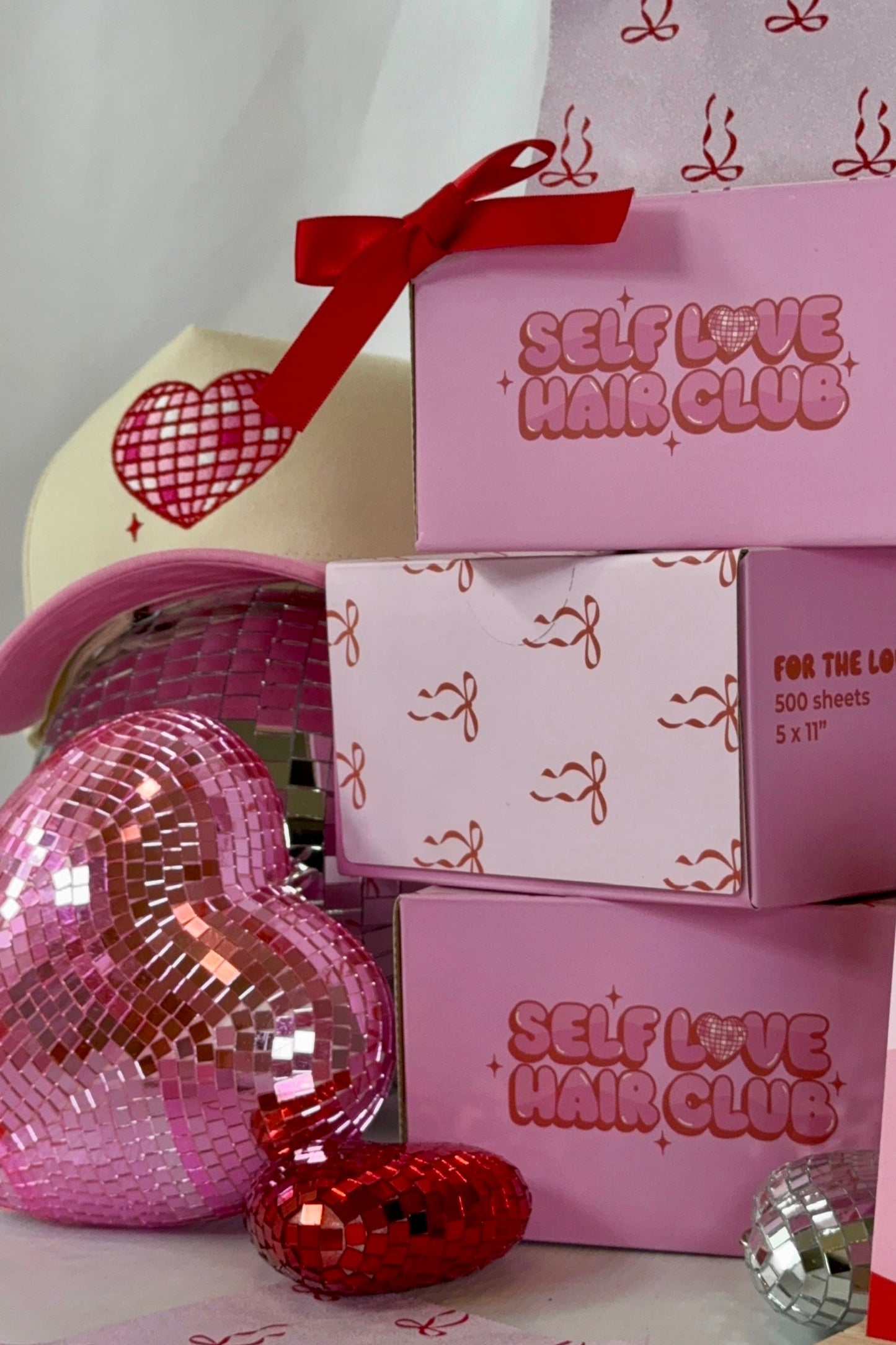 Pink Self Love Hair Club foil boxes with red bow design stacked beside a pink and cream hat surrounded by heart-shaped disco balls.