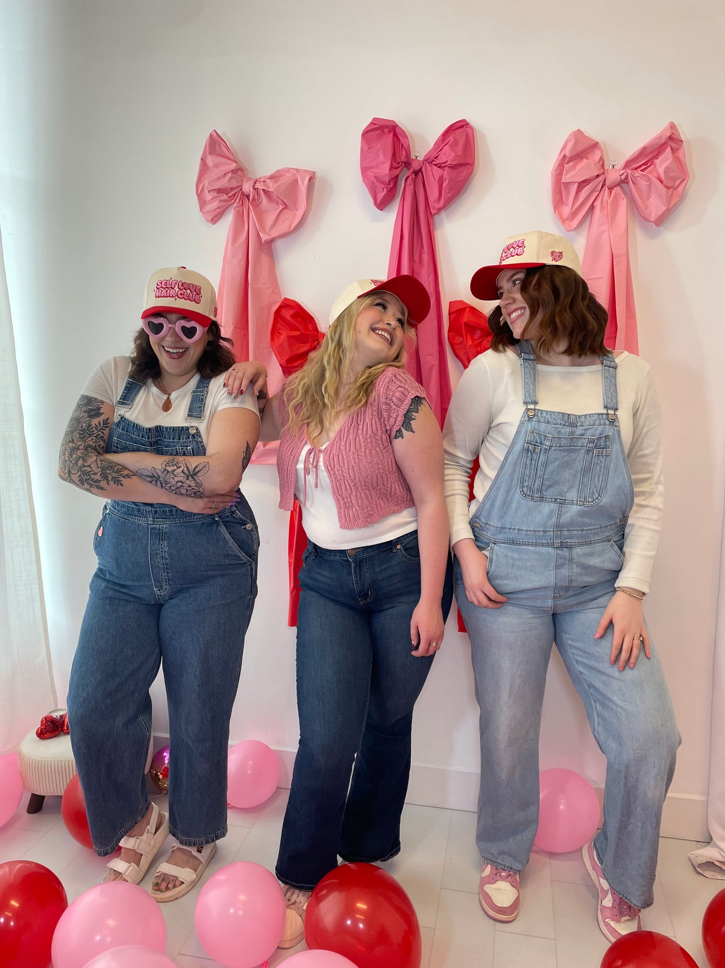 Three women posing together wearing Self Love Hair Club hats with balloons and ribbons on a white background