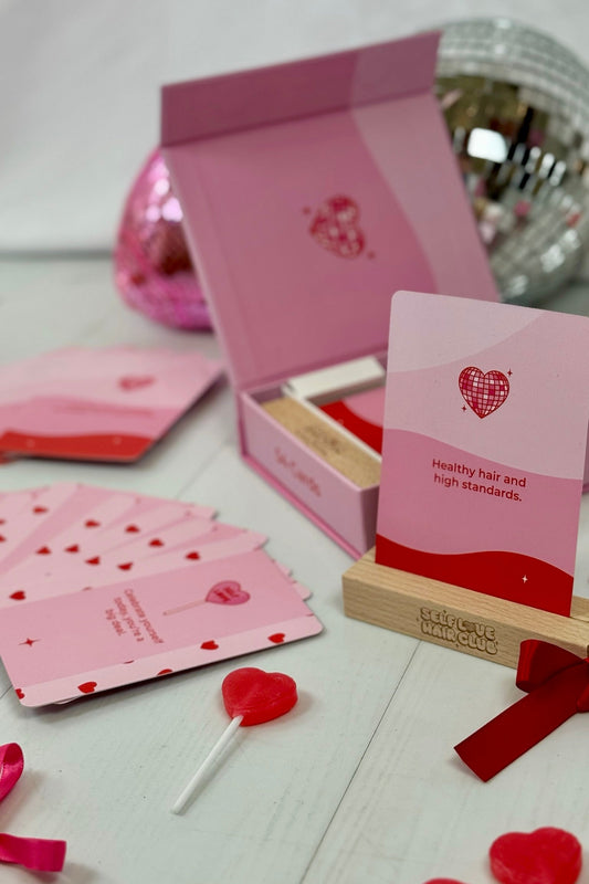 Self Love Hair Club affirmation card reading "Healthy hair and high standards" displayed on a wooden stand, with heart-themed cards, pink gift box, and disco balls in the background. 