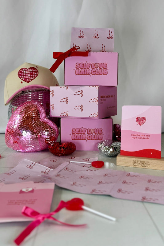 Self Love Hair Club foil boxes, affirmation card on wooden stand, red bow print foils, pink and cream trucker hat with disco heart, styled with heart-shaped disco balls. 