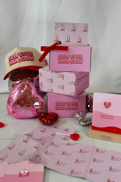 Collection of Self Love Hair Club products including foil boxes, affirmation card on a wooden display stand, and red with cream hat on a white background.