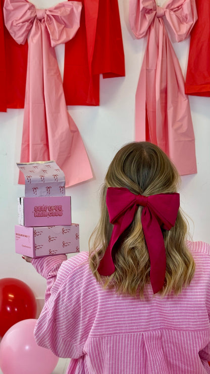 Half-up hairstyle with red bow holding Self Love Hair Club foil boxes in front of oversized fabric bows. 