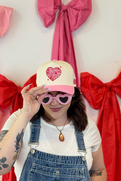 Person wearing a pink and cream Self Love Hair Club hat with red disco heart design, sunglasses, and overalls in front of red and pink bows.