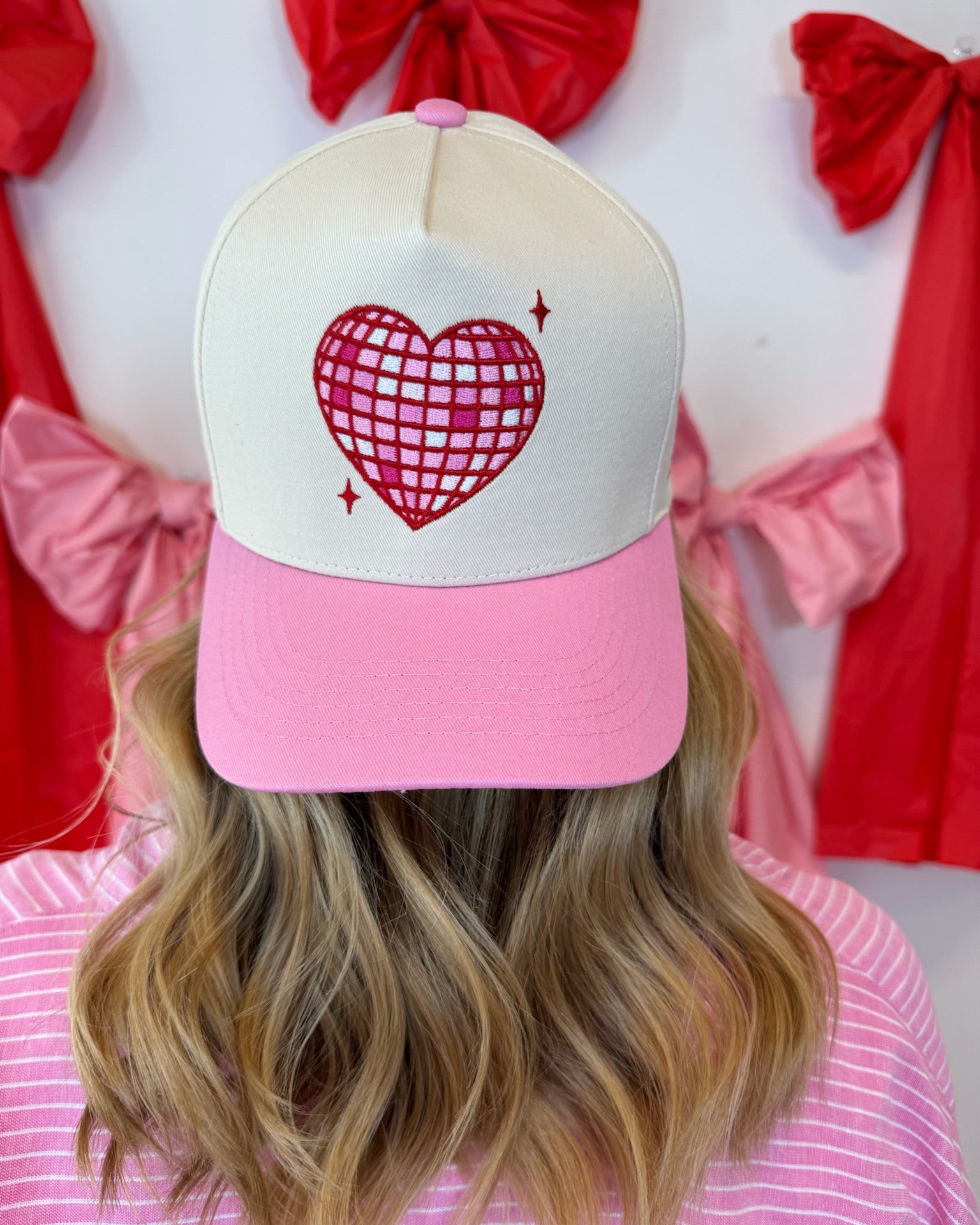 Blonde hair wearing a pink and cream Self Love Hair Club hat with a red disco heart design, standing in front of red bows.