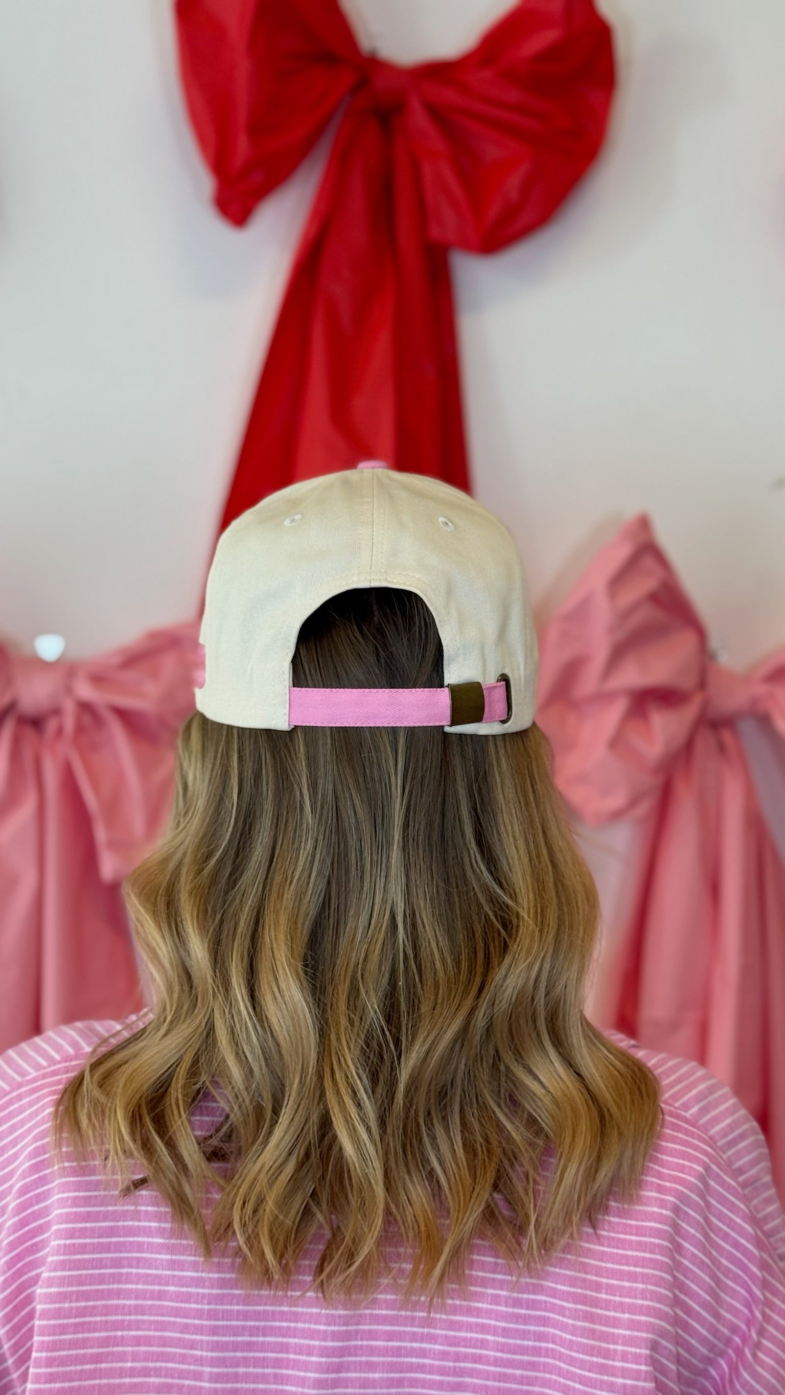 Person wearing a pink and cream Self Love Hair Club hat with a pink strap in front of pink and red bows.