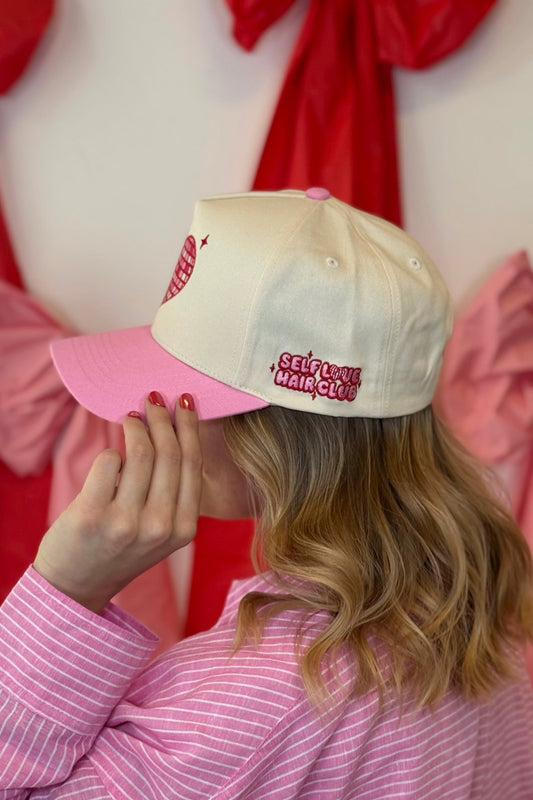 Person with blonde hair wearing a pink and cream Self Love Hair Club hat, standing against a red and white background.