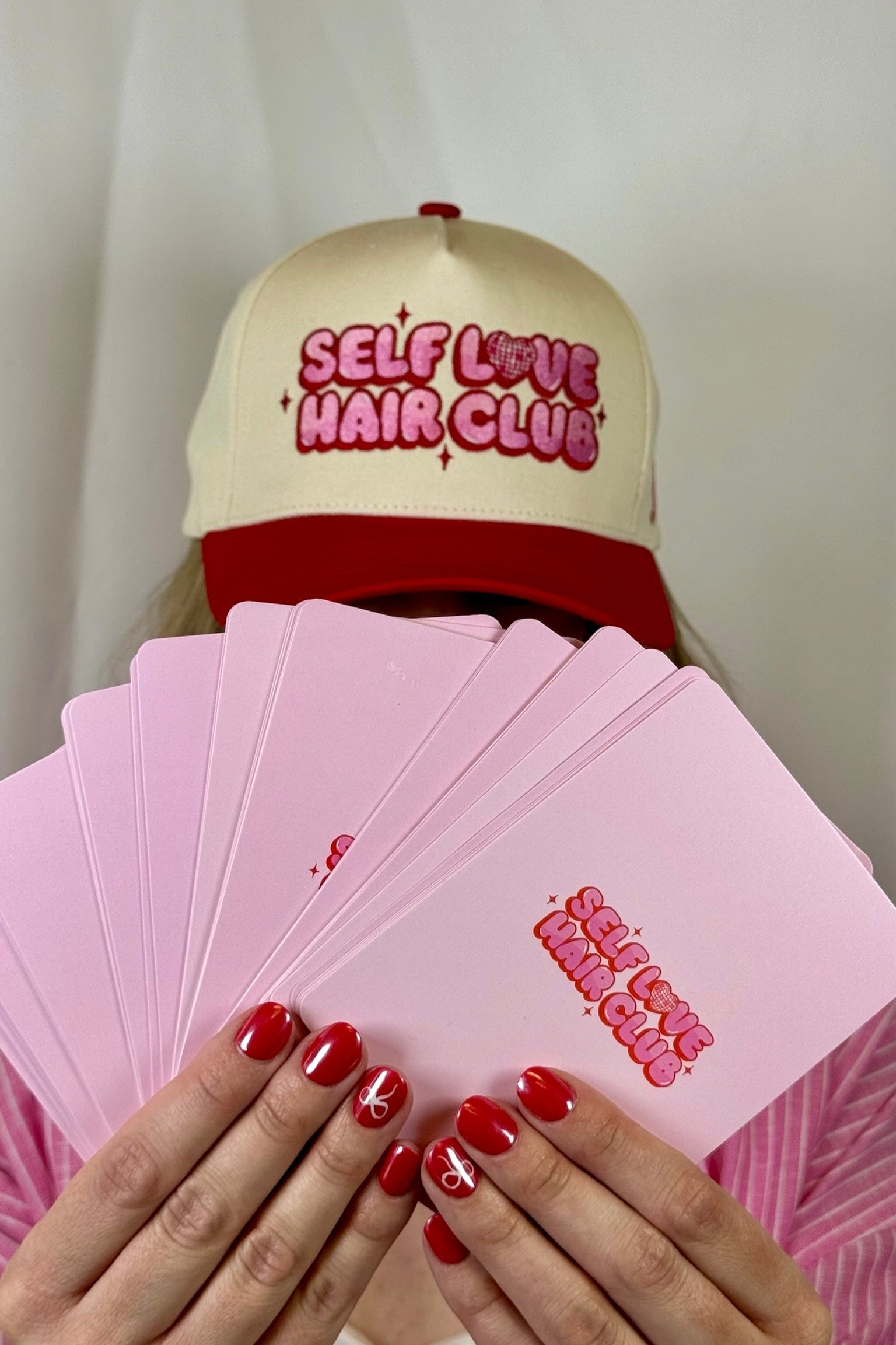 Person wearing a Self Love Hair Club hat and holding pink Self Love Hair Club affirmation cards.