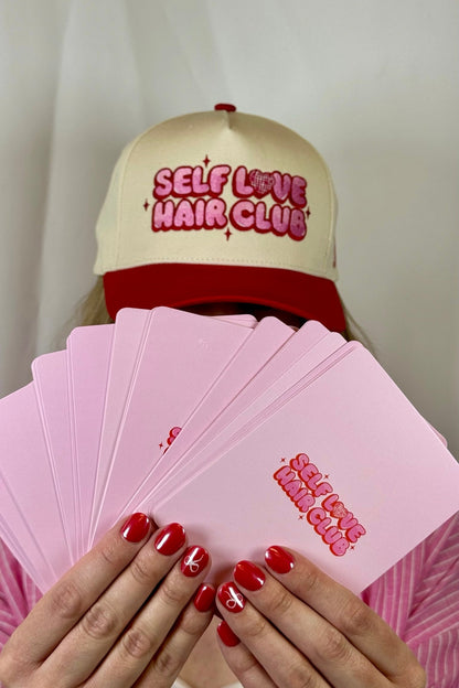 Person wearing a Self Love Hair Club hat and holding pink Self Love Hair Club affirmation cards.
