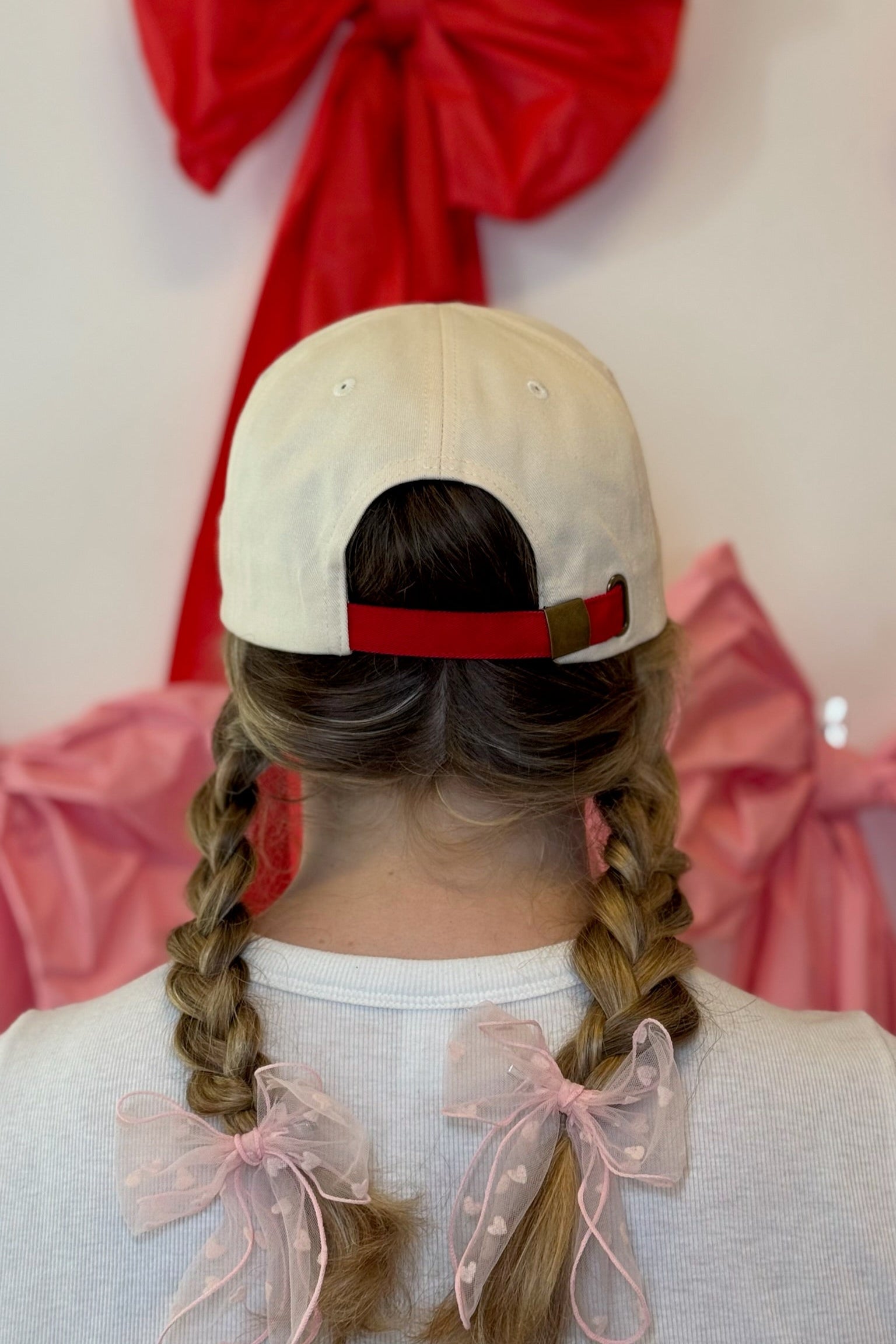Person wearing a Self Love Hair Club  hat with a red band, braided hair with bows, and a pink and red bow in the background.