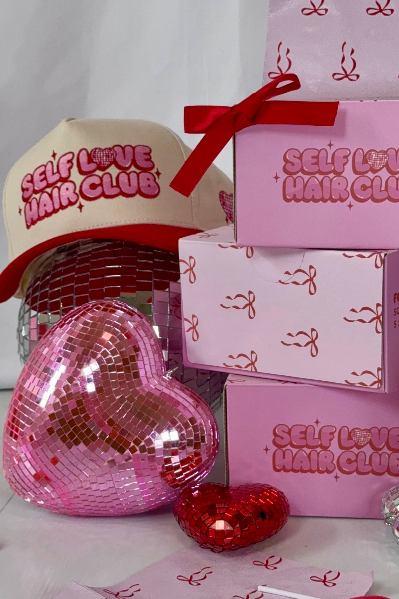 Pink Self Love Hair Club foil boxes with red bow design stacked beside a red and cream hat surrounded by heart-shaped disco balls.