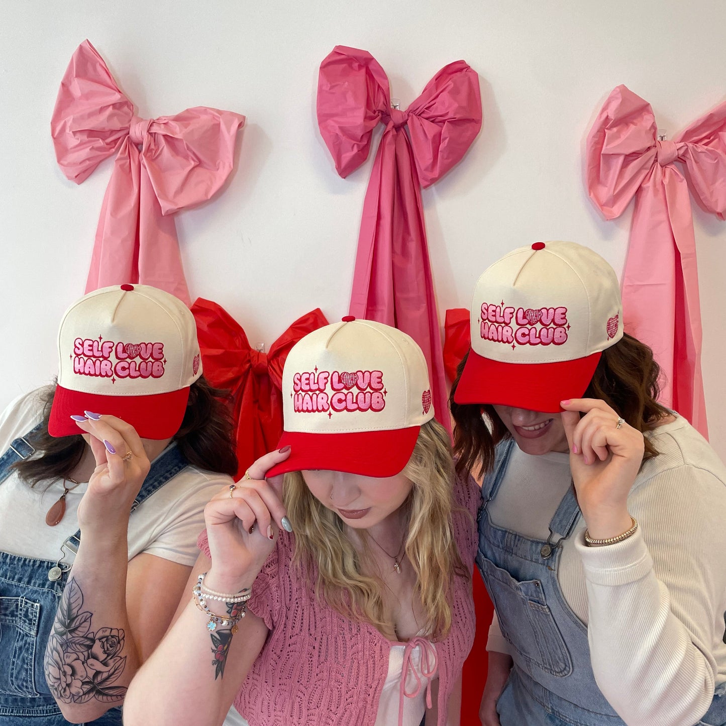 Three people wearing red and cream Self Love Hair Club hats with a white background and oversized fabric bows.
