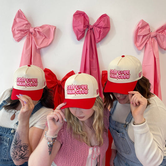 Three people wearing red and cream Self Love Hair Club hats with a white background and oversized fabric bows.