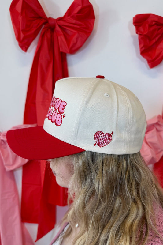 Person wearing a red and cream Self Love Hair Club hat, standing against a red and white background.